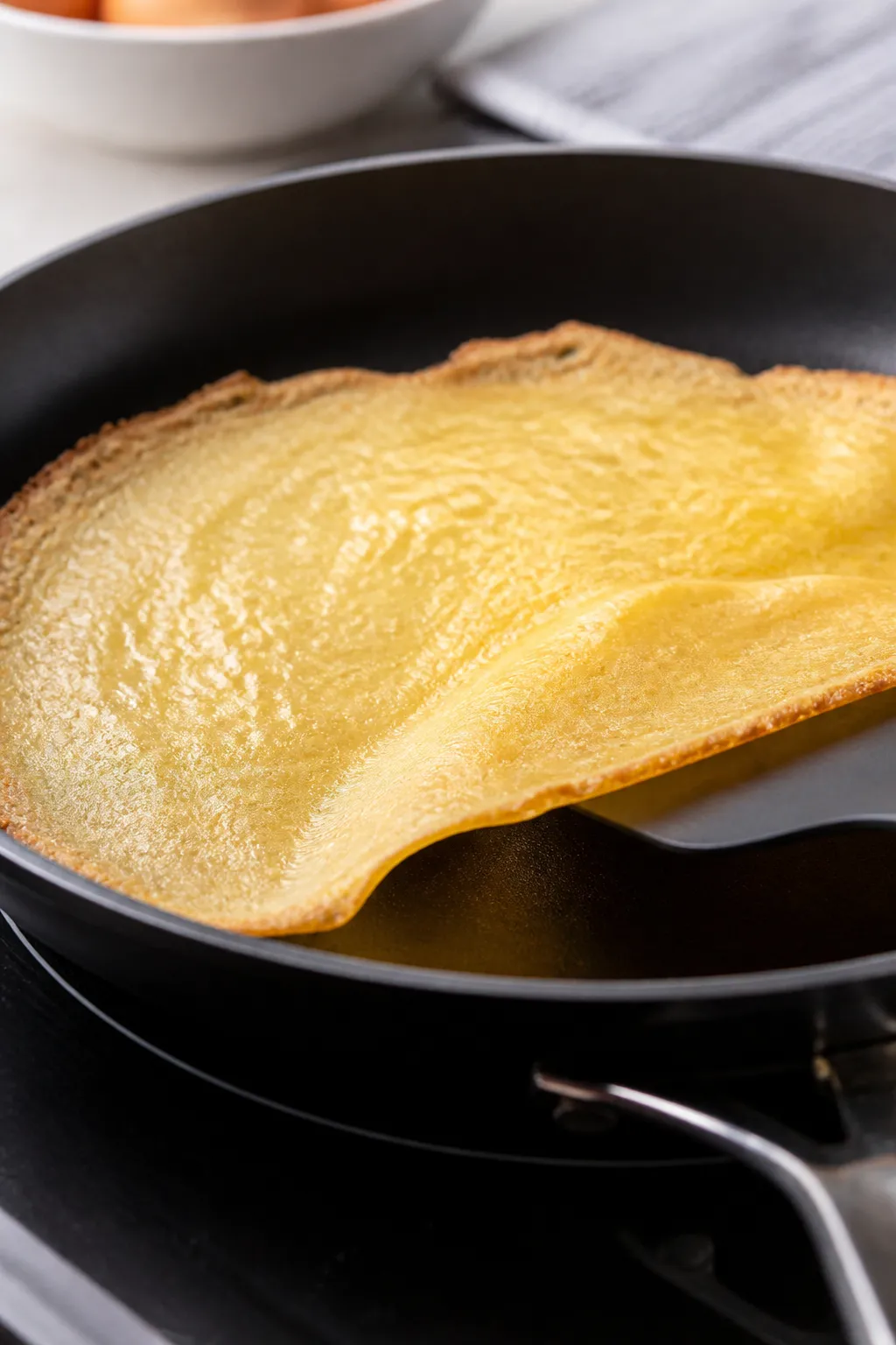 Cooking process close-up: A thin keto crepe in a nonstick skillet mid-cook, edges just set and light